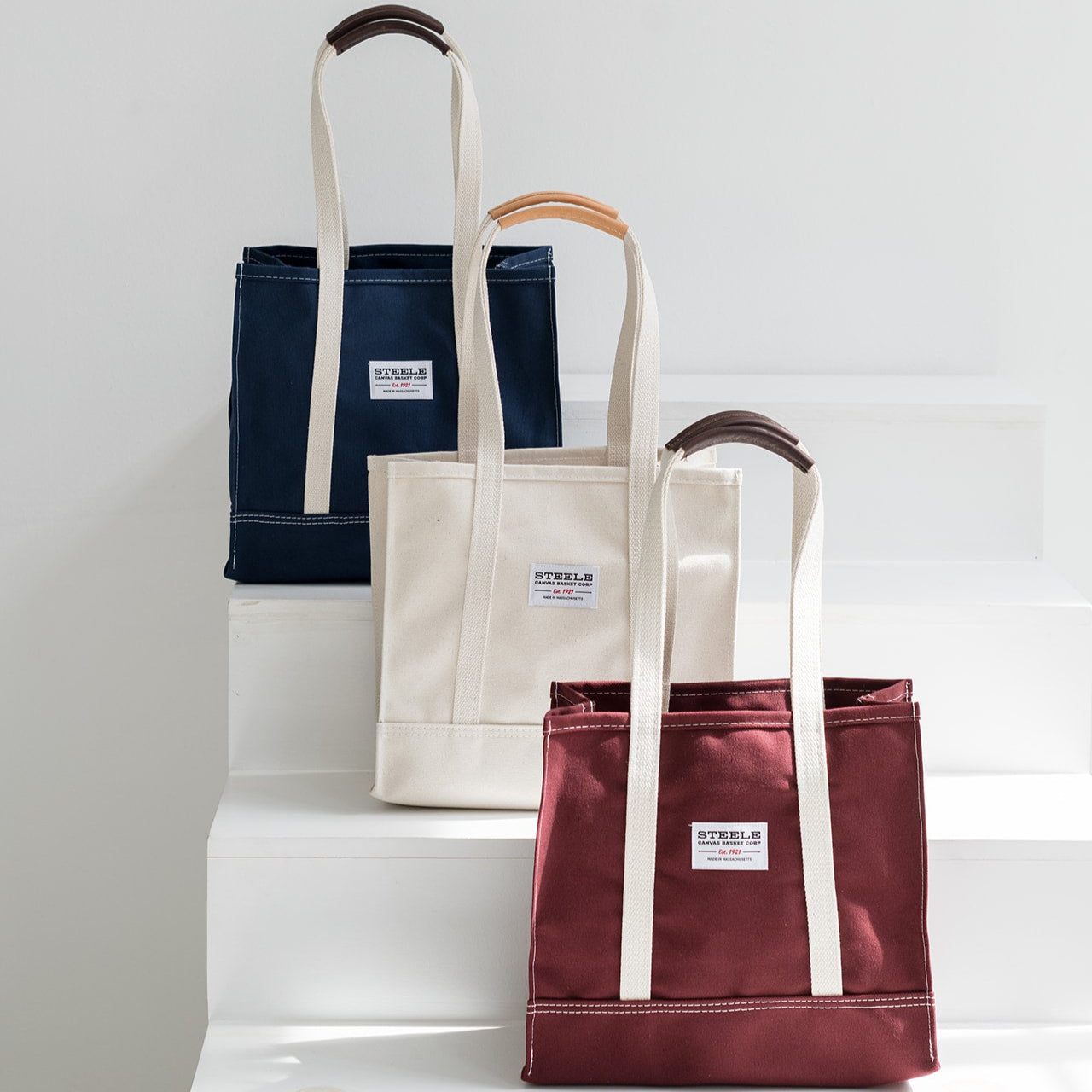 three tote bags in navy, beige, and red on a white surface with a light gray background.