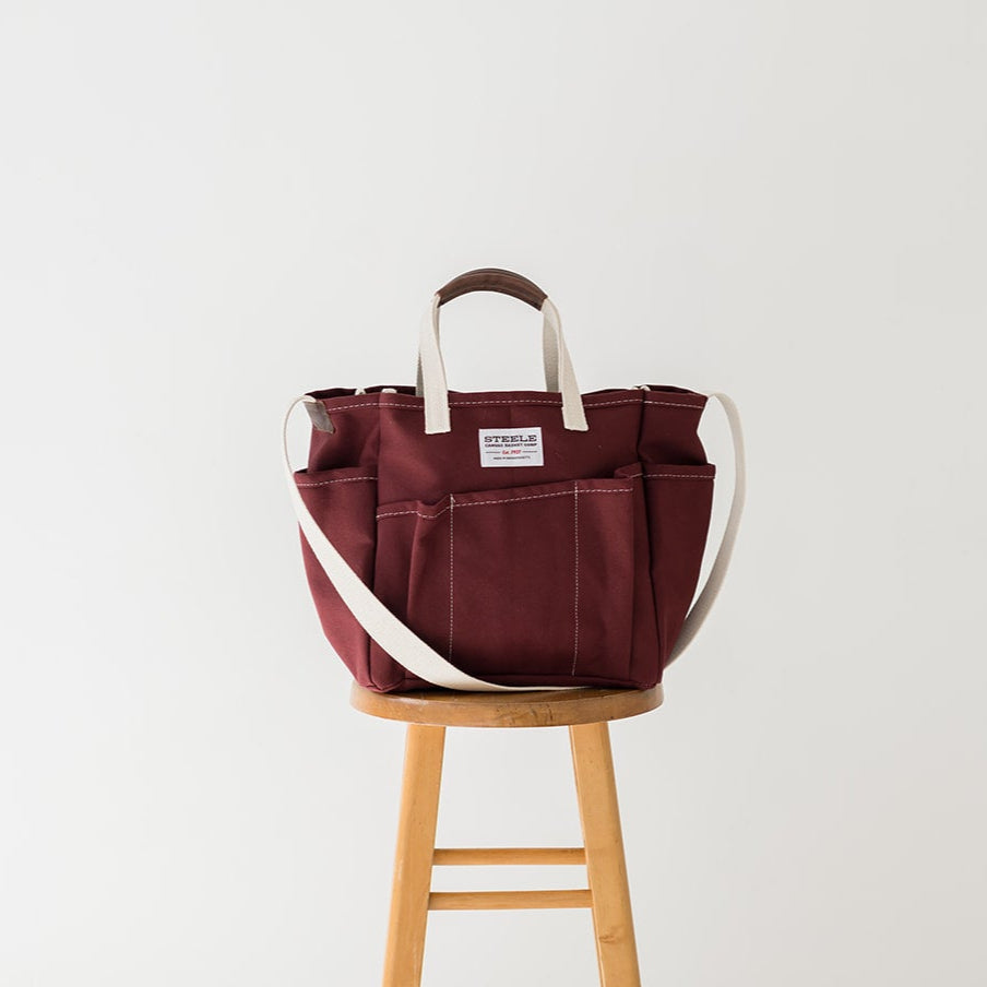 Maroon tote bag with white strap on a wooden stool against a white background