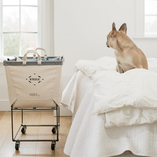 A natural canvas laundry truck on wheels, with a black metal frame, placed on a bed in a room with a dog sitting beside it.