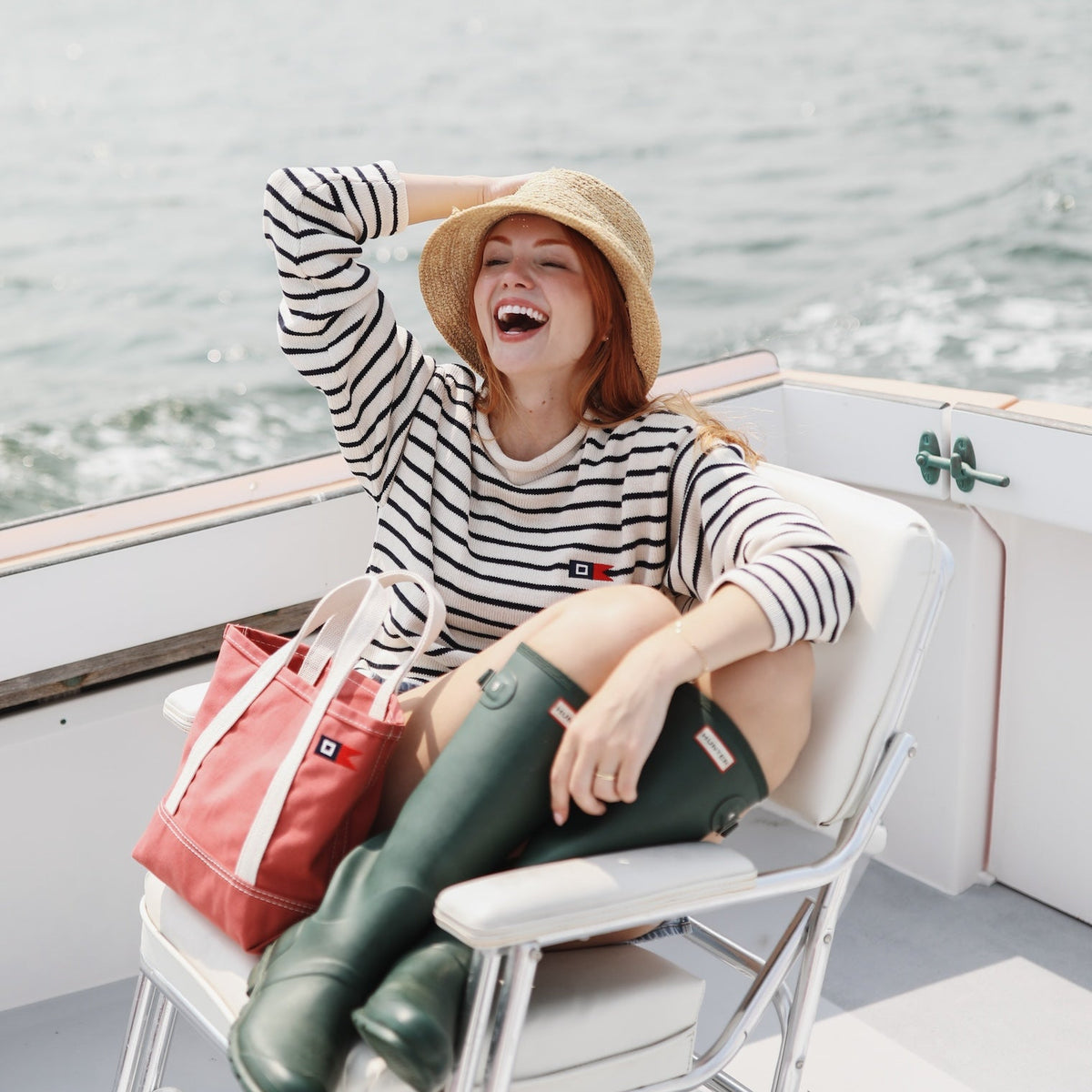 A woman laughing on a boat deck wearing a black and white striped long-sleeve top, a straw hat, dark green rain boots, and holding a red tote bag.