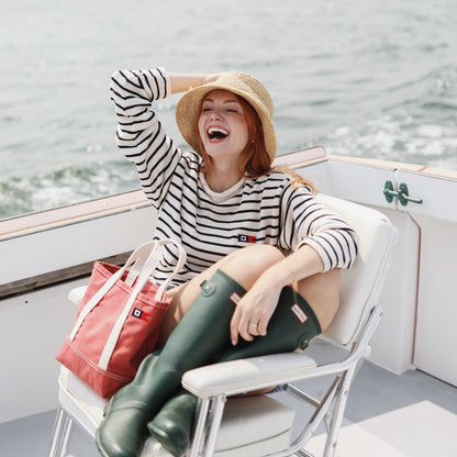 A woman laughing on a boat deck wearing a black and white striped long-sleeve top, a straw hat, dark green rain boots, and holding a red tote bag.