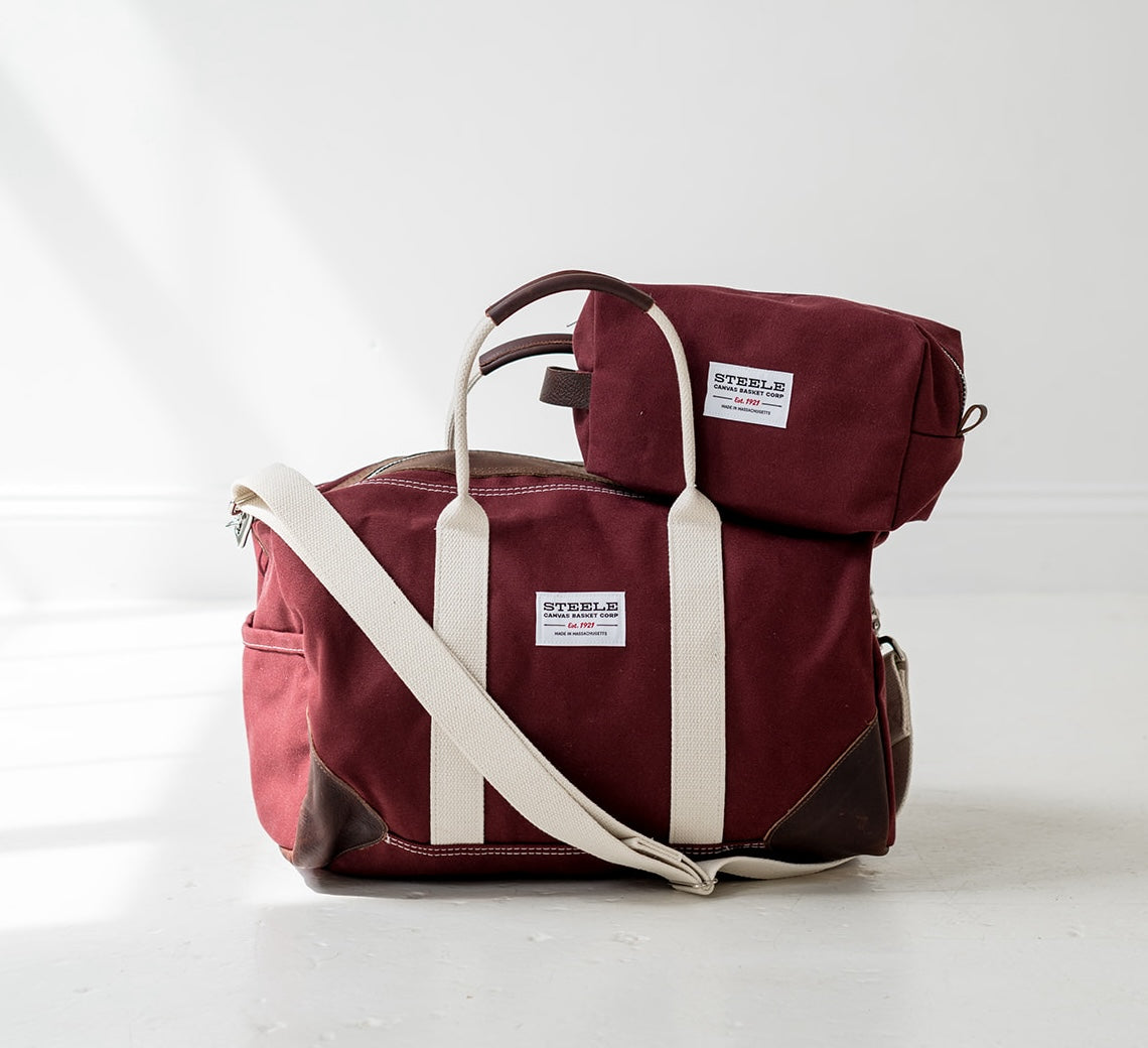Maroon duffel bag and dopp kit with white straps on a light gray background