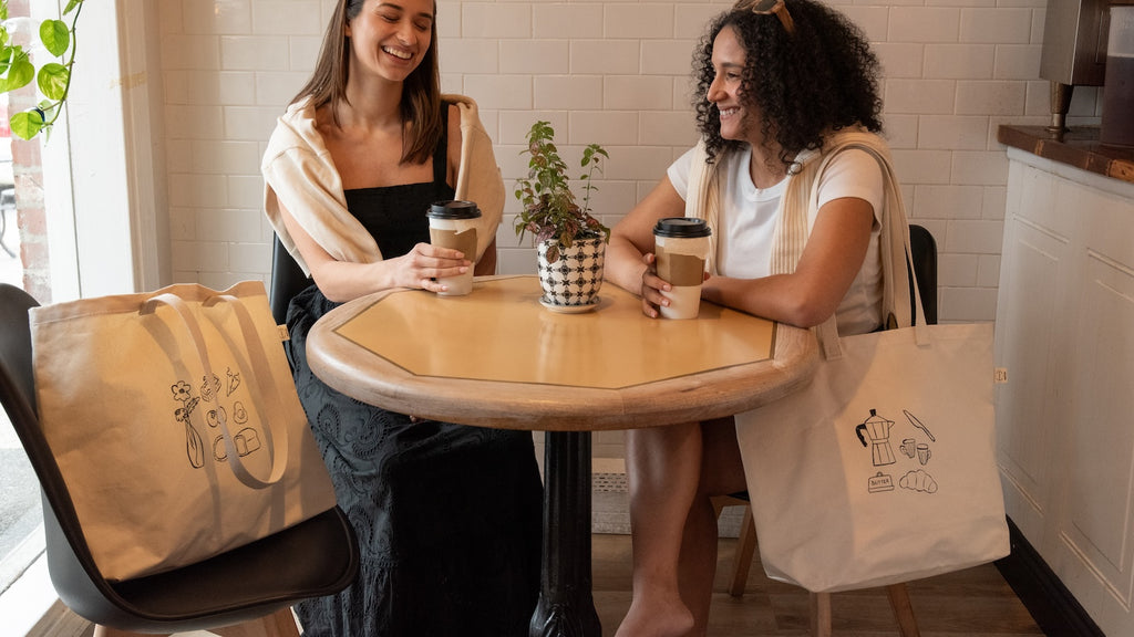 Two women sitting at a table with coffee cups and tote bags in a casual setting.
