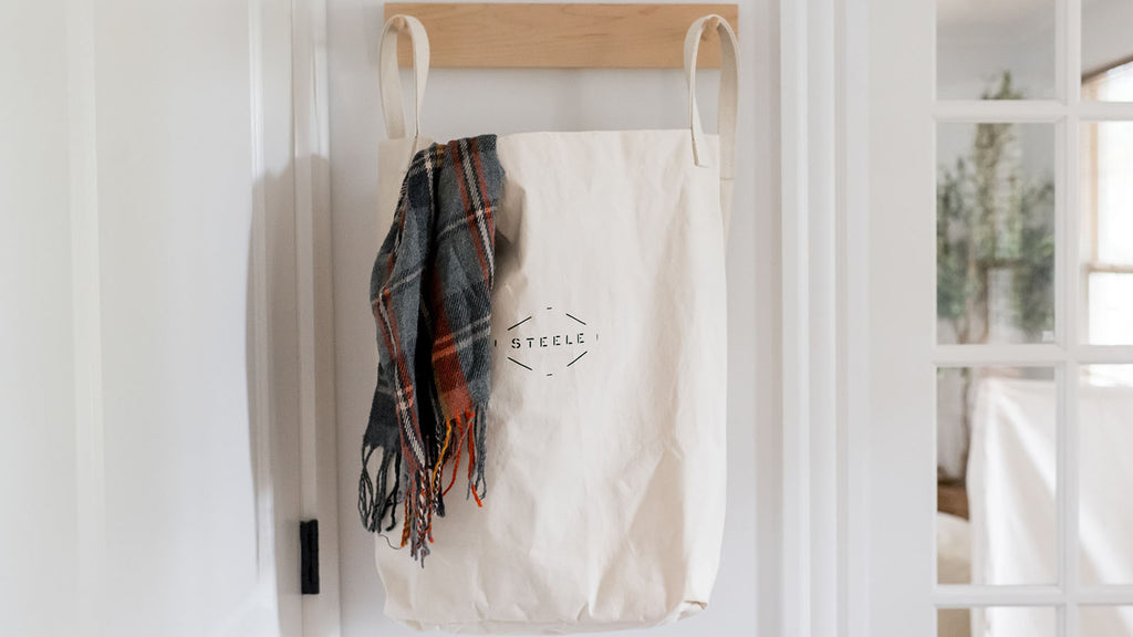 White tote bag with plaid scarf hanging on a wooden rack against a white wall.