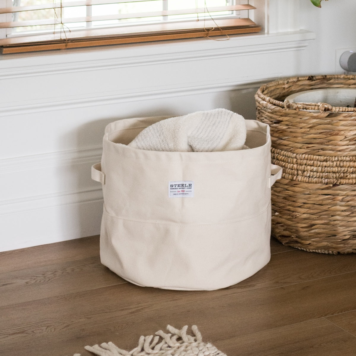 Beige storage bin with a towel inside, next to a woven basket on a wooden floor.