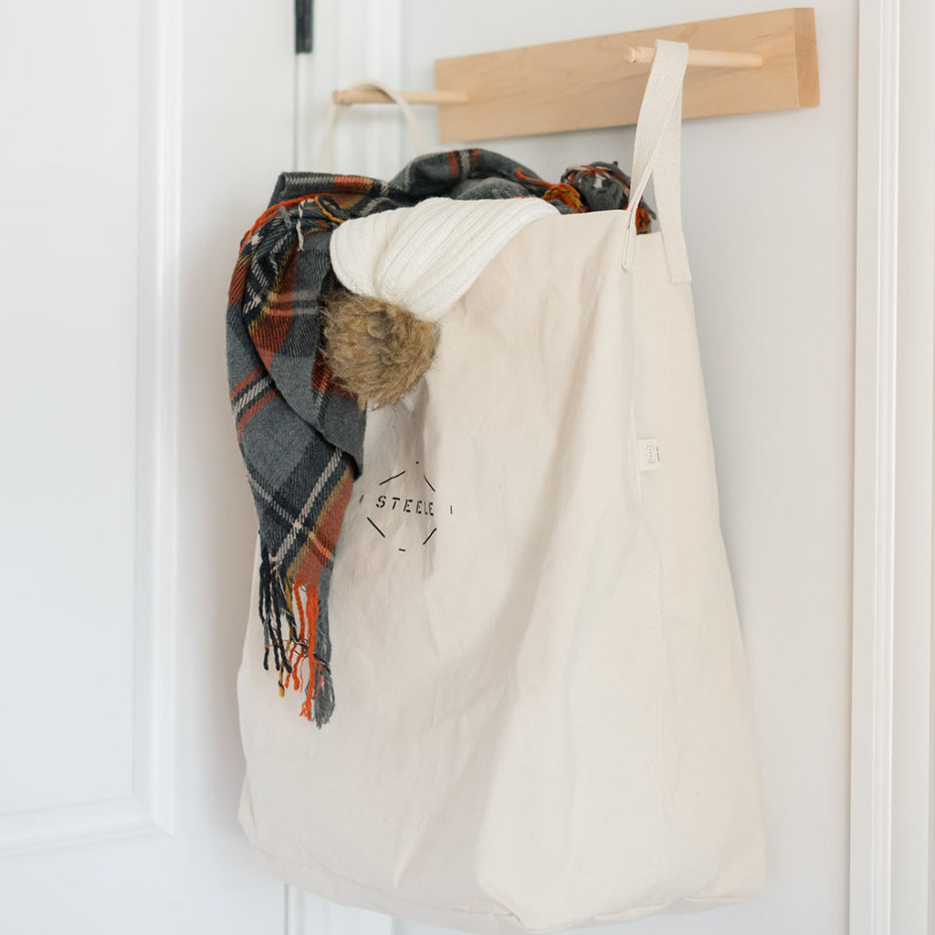White tote bag with plaid scarf hanging on a wooden rack against a white wall.