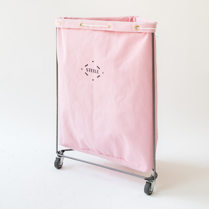 A pink canvas laundry hamper with a metal frame and caster wheels, featuring the brand name 'STEELE' printed in black on the front.