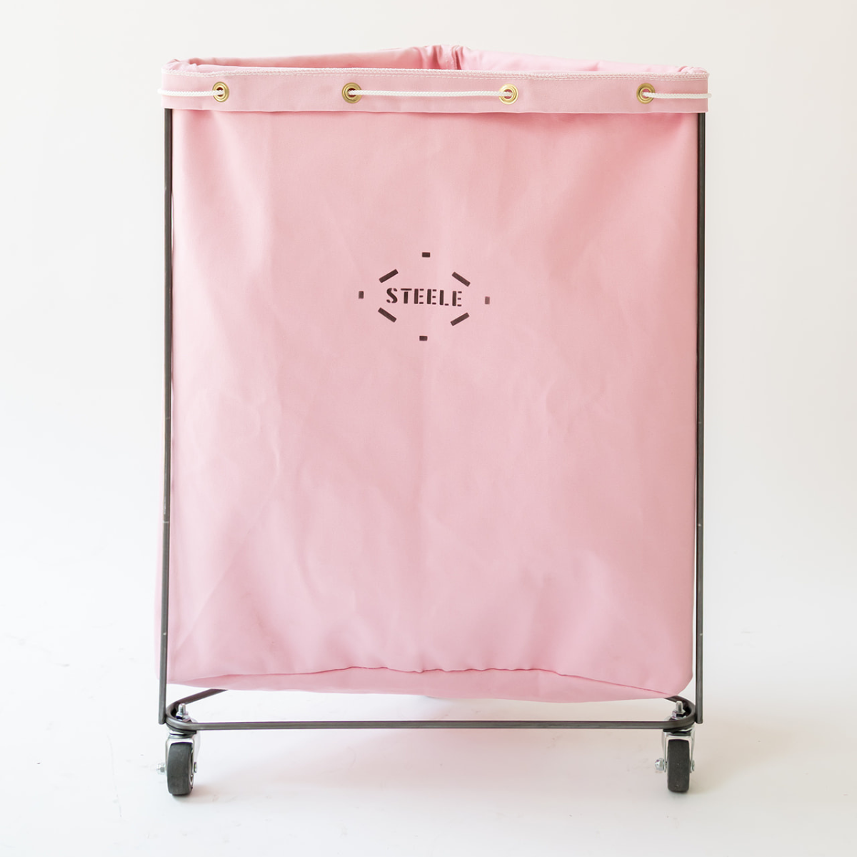 A pink canvas laundry hamper bag with a black metal frame and wheels, featuring the brand name 'STEELE' printed in black in the center. The top of the bag has gold grommets.
