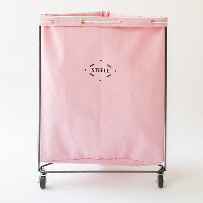 A pink canvas laundry hamper bag with a black metal frame and wheels, featuring the brand name 'STEELE' printed in black in the center. The top of the bag has gold grommets.