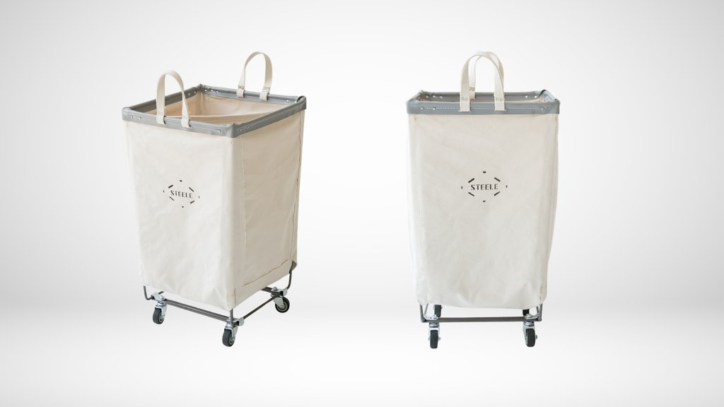 Two fabric laundry hamper carts shown on a light gray studio background. Each cart has a cream canvas body, gray trim, white top handles, and a black metal wheeled frame.