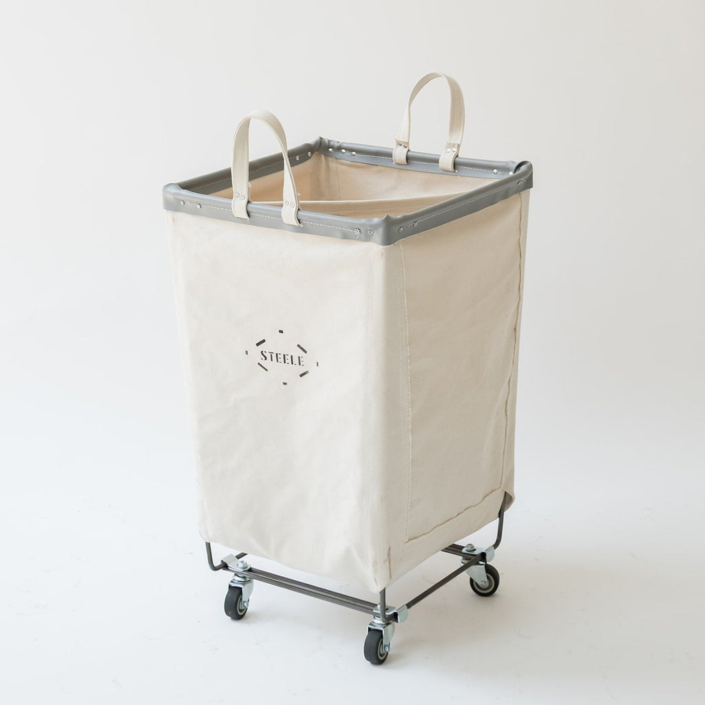 Two fabric laundry hamper carts shown on a light gray studio background. Each cart has a cream canvas body, gray trim, white top handles, and a black metal wheeled frame.